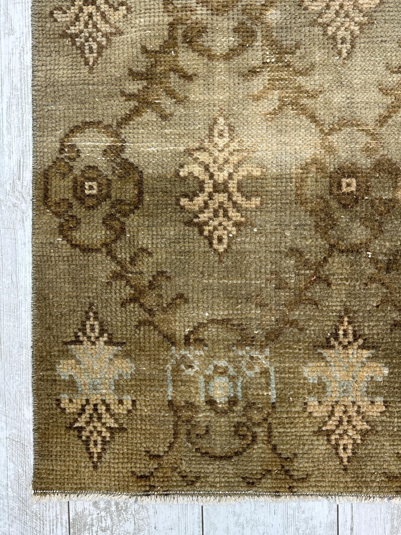 May include: A woven rug featuring a muted olive green and brown color palette with a repeating geometric pattern of stylized floral motifs and interconnected diamond shapes. The texture appears dense and low-pile with a fringed edge at the bottom.