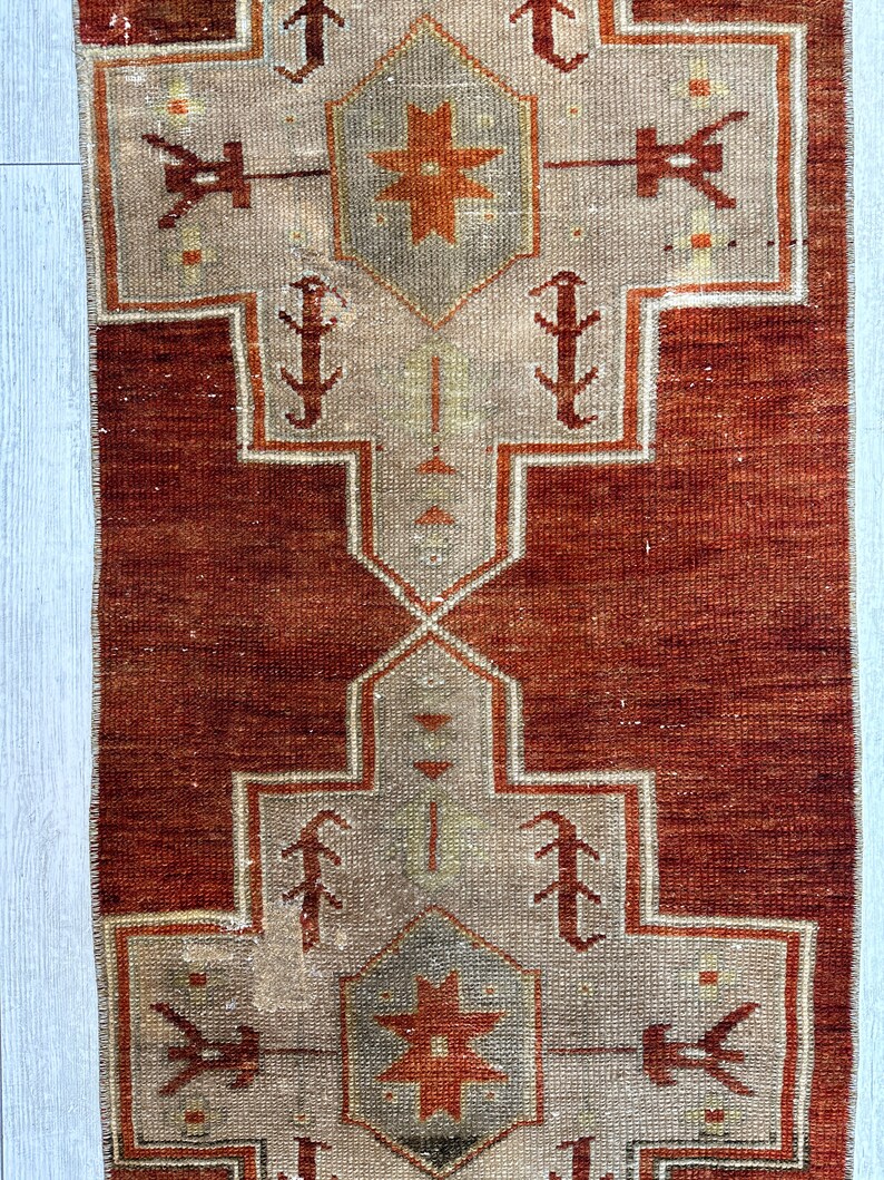 May include: A vintage Turkish runner rug with a red background and a beige pattern. The pattern features a central star with geometric shapes and stylized floral motifs.