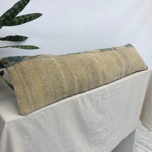 Puede incluir: Un cojín lumbar largo y rectangular con una tela beige tejida y texturizada. El cojín muestra patrones geométricos en azul y gris descoloridos a lo largo del borde superior, exhibiendo un aspecto vintage desgastado con hilos visibles.