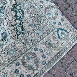 Puede incluir: Una alfombra vintage con fondo crema y un patr&oacute;n geom&eacute;trico azul y verde. La alfombra tiene un borde deshilachado y est&aacute; sobre una superficie de ladrillo.