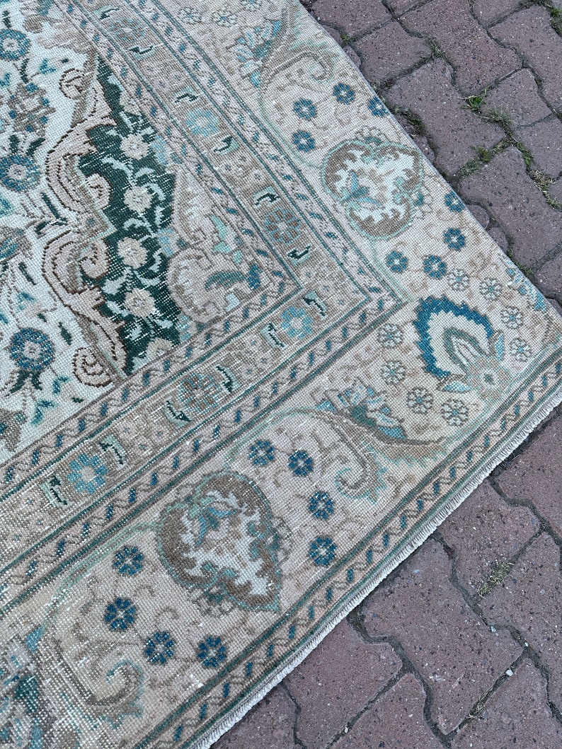 Puede incluir: Una alfombra vintage con fondo crema y un patr&oacute;n geom&eacute;trico azul y verde. La alfombra tiene un borde deshilachado y est&aacute; sobre una superficie de ladrillo.