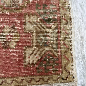 Puede incluir: Primer plano de una alfombra vintage con un aspecto desgastado y descolorido. La alfombra presenta un dise&ntilde;o central en tonos rojos, marrones y beige, con patrones geom&eacute;tricos y un borde con flecos. La textura de la alfombra es visible, mostrando su construcci&oacute;n tejida.