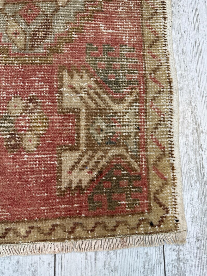 Puede incluir: Primer plano de una alfombra vintage con un aspecto desgastado y descolorido. La alfombra presenta un dise&ntilde;o central en tonos rojos, marrones y beige, con patrones geom&eacute;tricos y un borde con flecos. La textura de la alfombra es visible, mostrando su construcci&oacute;n tejida.