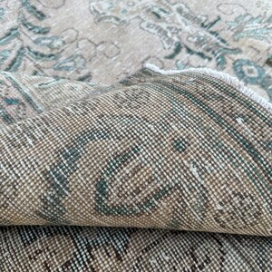 Puede incluir: Primer plano del reverso de una alfombra beige con un patr&oacute;n descolorido azul y marr&oacute;n. La alfombra est&aacute; doblada por la mitad, mostrando la textura tejida.