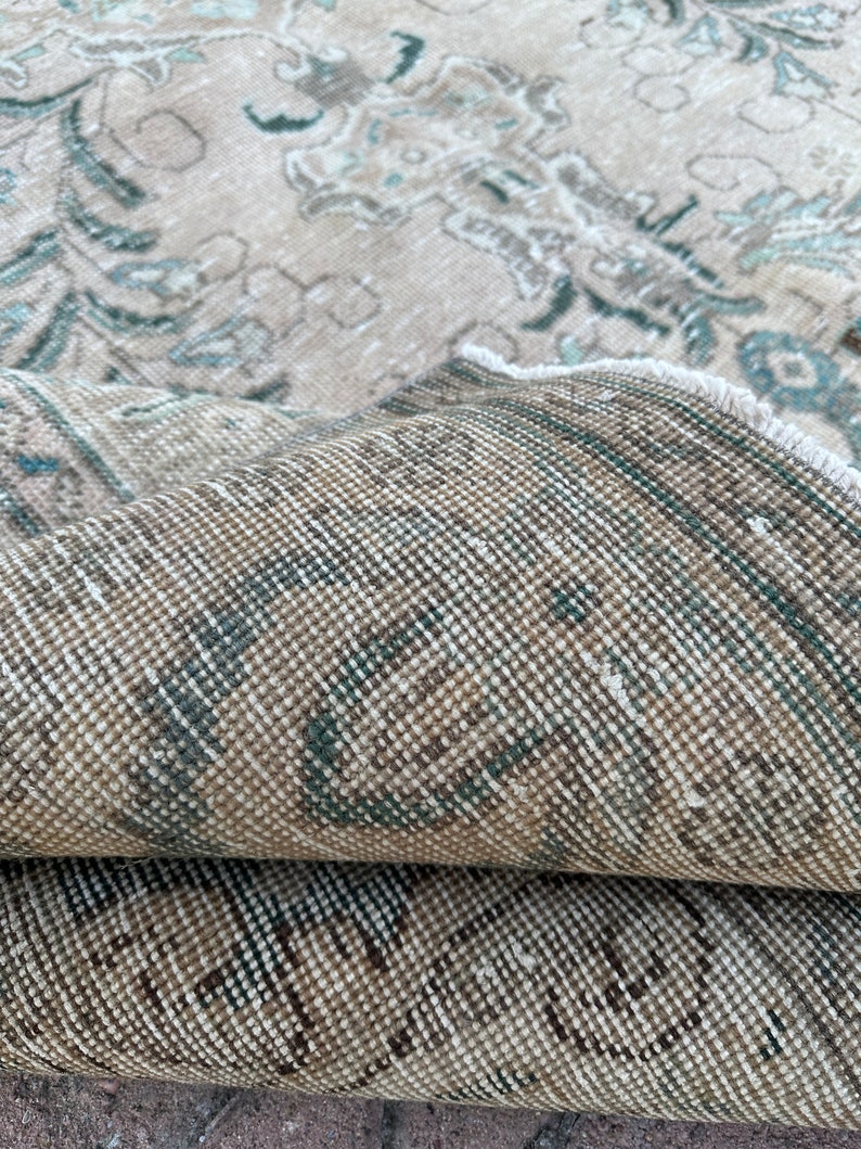 Puede incluir: Primer plano del reverso de una alfombra beige con un patr&oacute;n descolorido azul y marr&oacute;n. La alfombra est&aacute; doblada por la mitad, mostrando la textura tejida.