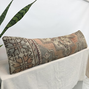 Puede incluir: Un cojín lumbar rectangular largo con una funda de tejido con un patrón geométrico descolorido en tonos apagados de melocotón, verde salvia, beige y marrón. La tela tiene una apariencia texturizada y desgastada con motivos tribales.