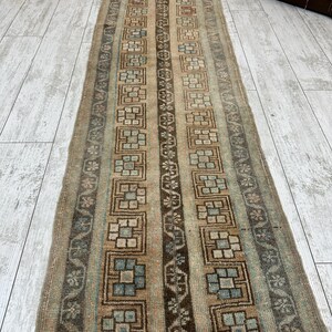 May include: A rectangular, vintage-style rug with a repeating geometric pattern in shades of brown, beige, and blue. The rug features a central column of dark brown and beige squares, framed by vertical stripes and floral motifs. The rug is placed on a light-colored wooden floor.