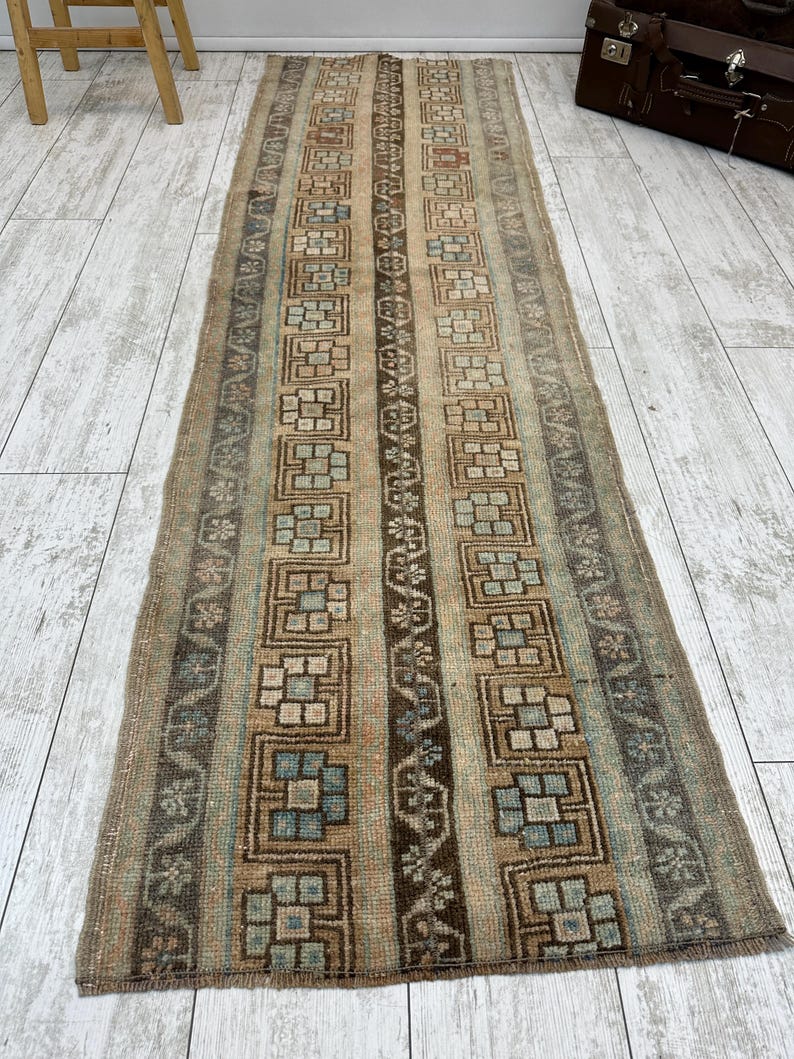 May include: A rectangular, vintage-style rug with a repeating geometric pattern in shades of brown, beige, and blue. The rug features a central column of dark brown and beige squares, framed by vertical stripes and floral motifs. The rug is placed on a light-colored wooden floor.