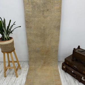 May include: A light beige runner rug with a subtle pattern and frayed edges. The rug is hanging on a white wall and is photographed against a white wood floor.
