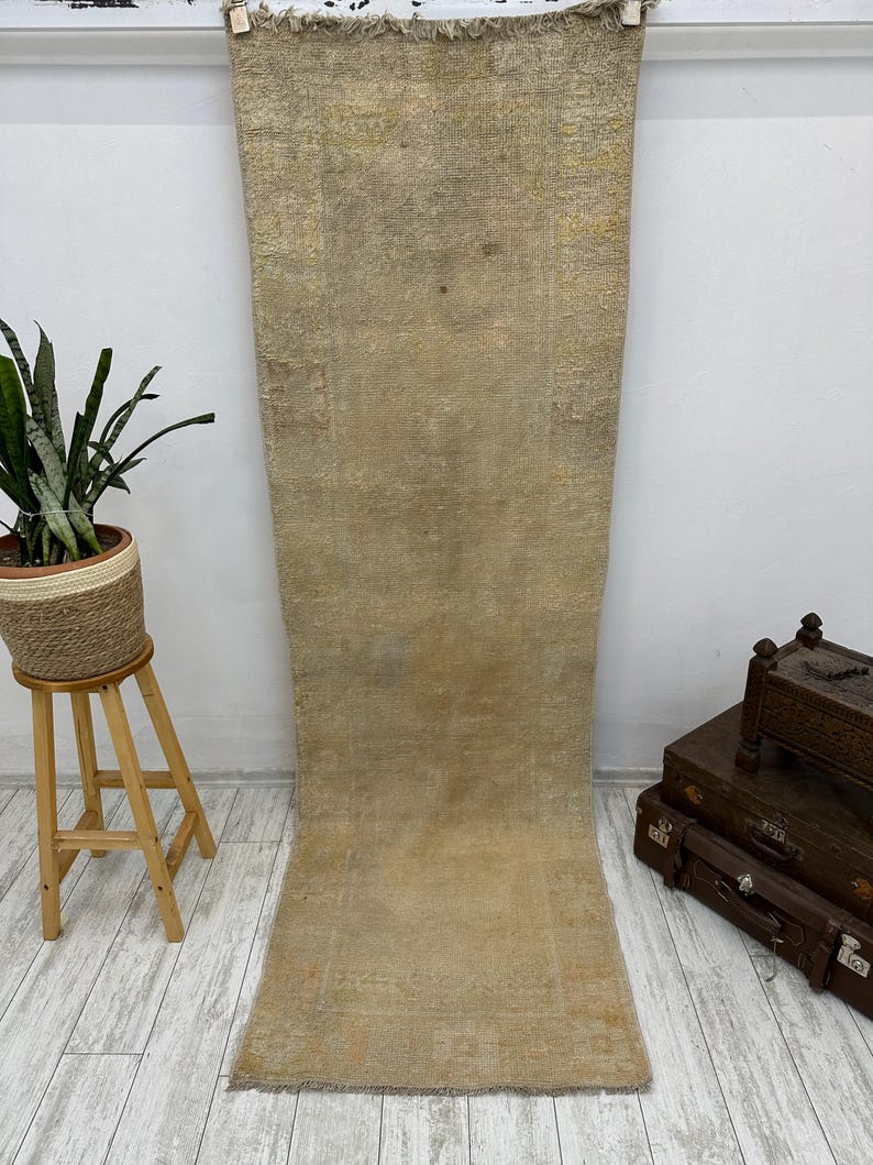 May include: A light beige runner rug with a subtle pattern and frayed edges. The rug is hanging on a white wall and is photographed against a white wood floor.