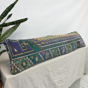 May include: A long, rectangular lumbar pillow featuring a woven textile cover with a complex geometric pattern in shades of deep purple, forest green, tan, and cream. The fabric displays a textured, knotted appearance with a visible zipper closure.