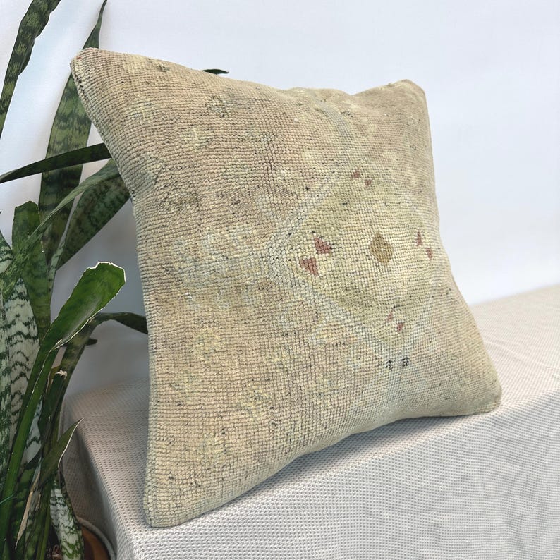 May include: A square throw pillow featuring a textured, woven surface in muted beige and tan tones. The fabric displays a faded, geometric diamond pattern in the center with subtle accents of soft pink and brown throughout the weave.