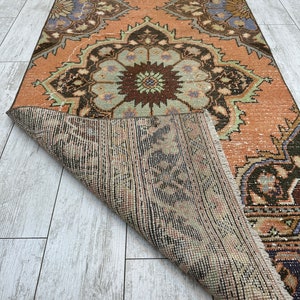 May include: A vintage rug with a peach background and intricate floral patterns in shades of brown, green, and blue. The rug is folded in half, revealing the woven backing.