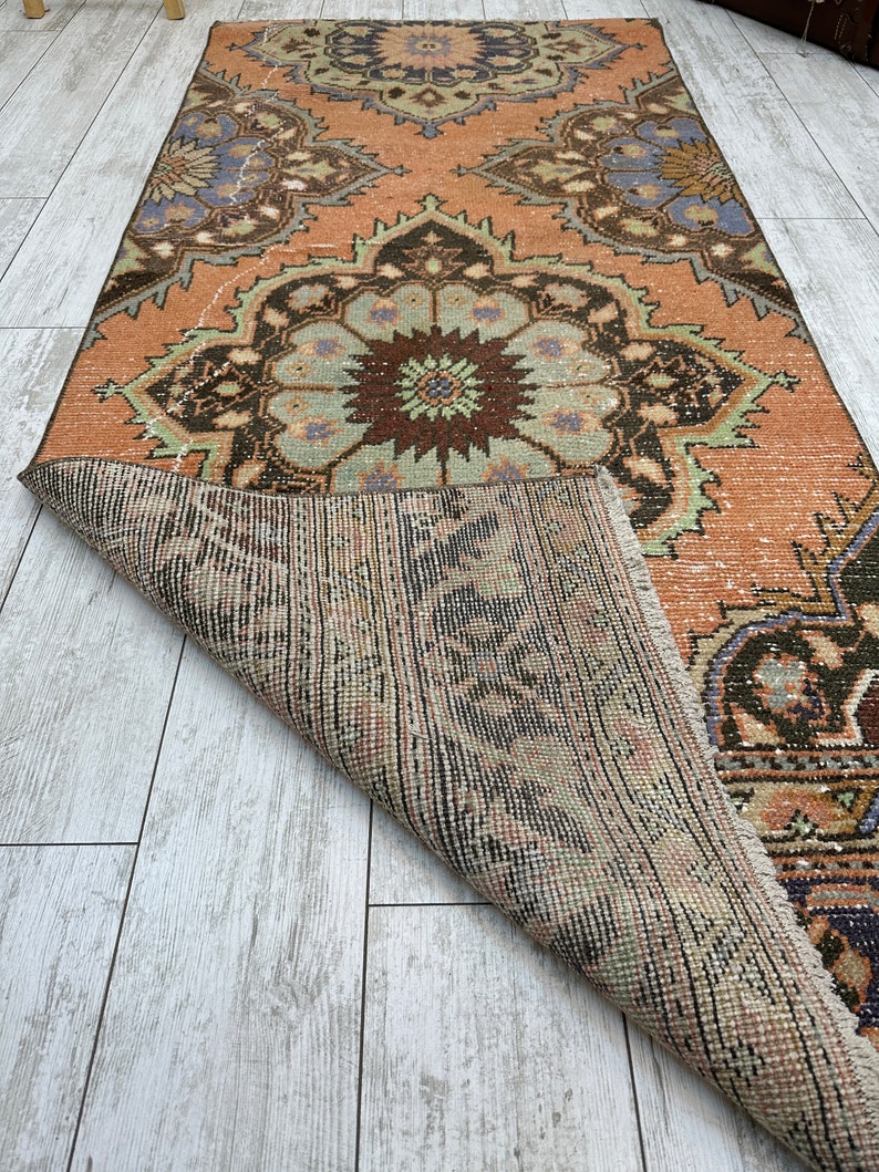May include: A vintage rug with a peach background and intricate floral patterns in shades of brown, green, and blue. The rug is folded in half, revealing the woven backing.
