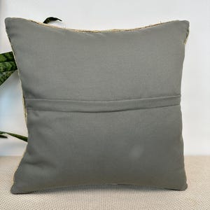 May include: A square throw pillow featuring a solid, matte gray fabric back panel with a horizontal seam running across the center. The pillow has a visible zipper closure along the top edge and a textured, woven border visible on the sides.