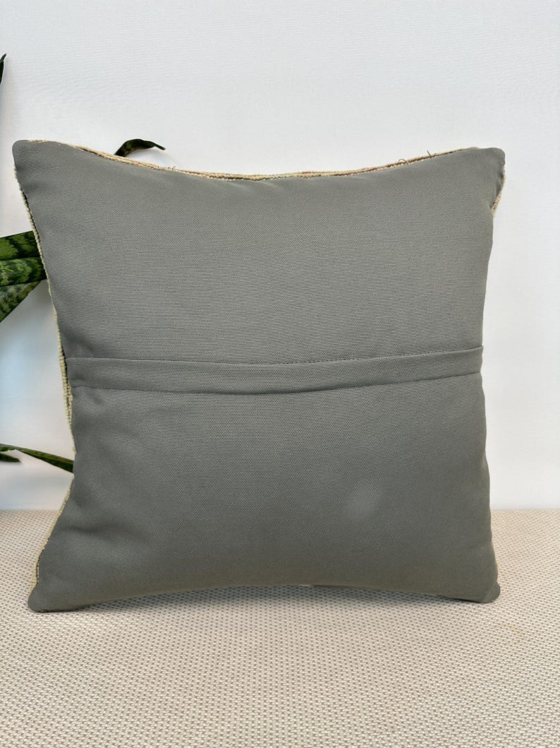 May include: A square throw pillow featuring a solid, matte gray fabric back panel with a horizontal seam running across the center. The pillow has a visible zipper closure along the top edge and a textured, woven border visible on the sides.