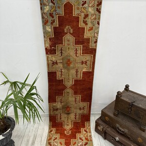 May include: A vintage Turkish runner rug with a red background and a geometric pattern in shades of beige, blue, and orange. The rug has a fringe border.