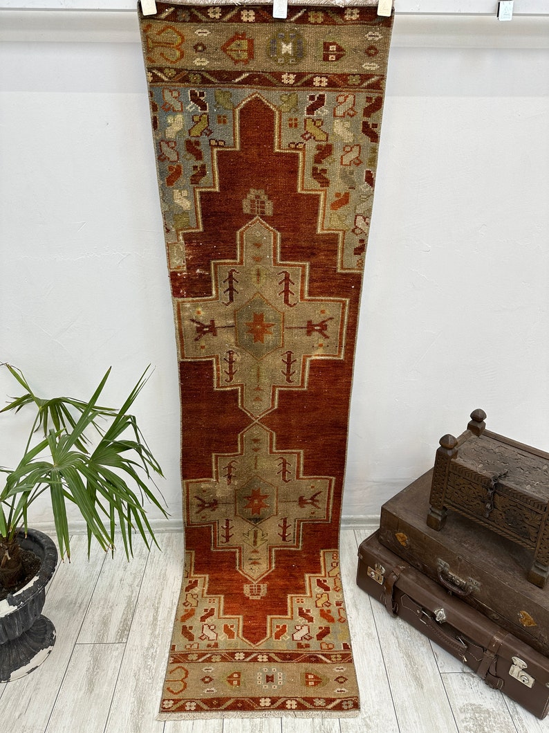 May include: A vintage Turkish runner rug with a red background and a geometric pattern in shades of beige, blue, and orange. The rug has a fringe border.