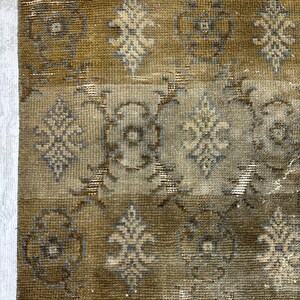 May include: A rectangular area rug featuring a muted olive green and brown color palette with a repeating pattern of stylized floral motifs and geometric shapes in light gray and beige. The rug has a short pile texture and a fringed top edge.