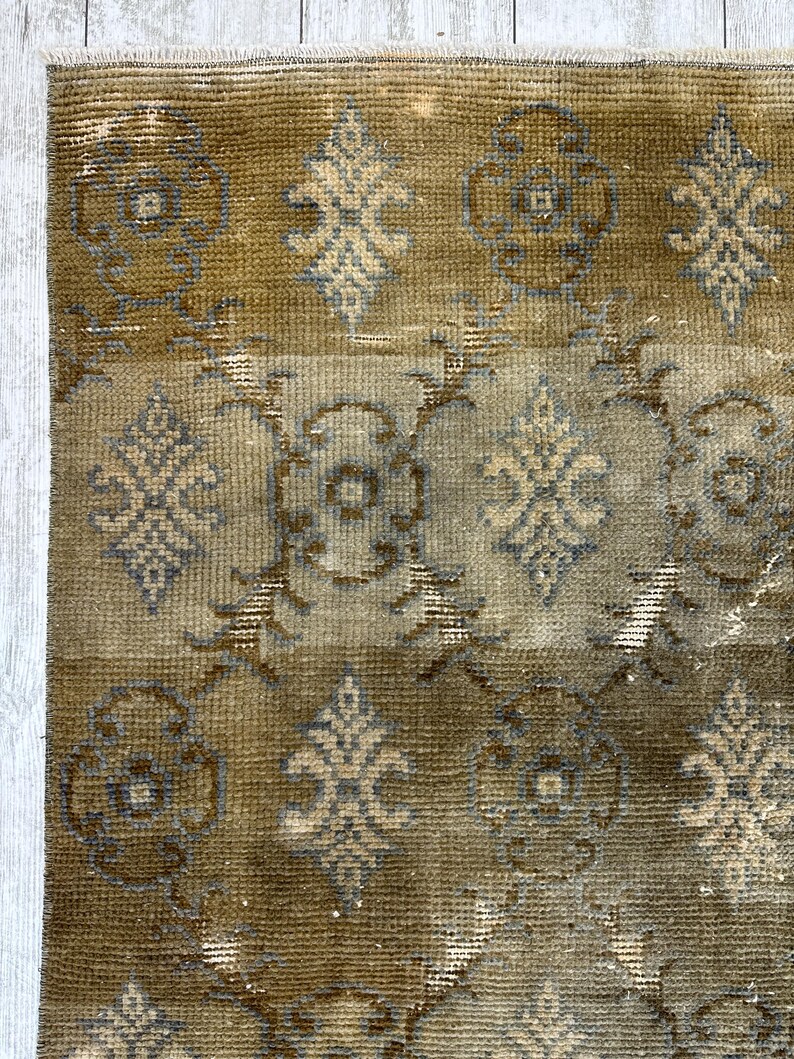 May include: A rectangular area rug featuring a muted olive green and brown color palette with a repeating pattern of stylized floral motifs and geometric shapes in light gray and beige. The rug has a short pile texture and a fringed top edge.