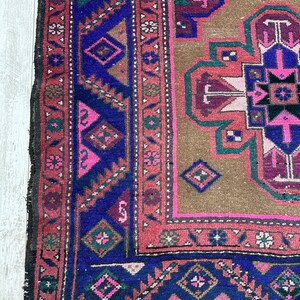 May include: A vintage rug with a brown background and a central medallion in shades of pink, purple, and blue. The rug has a geometric pattern with intricate details and a fringed edge.