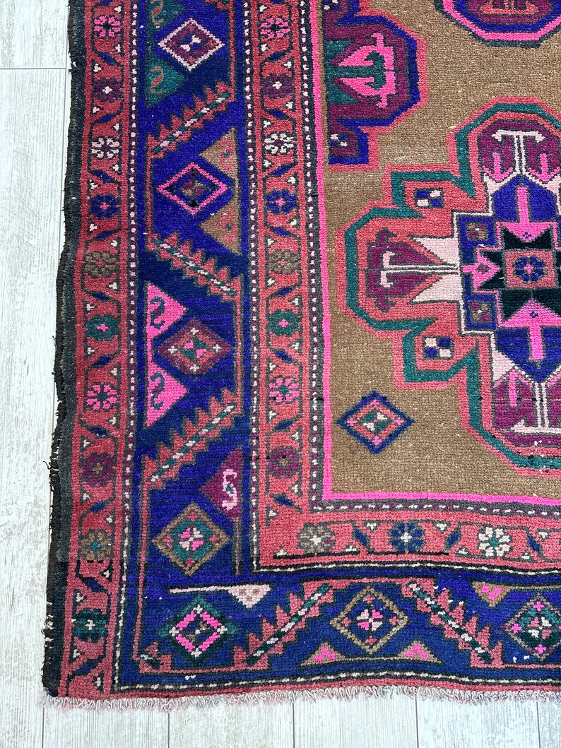 May include: A vintage rug with a brown background and a central medallion in shades of pink, purple, and blue. The rug has a geometric pattern with intricate details and a fringed edge.