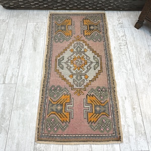 May include: A rectangular area rug featuring a dusty pink background with a central diamond motif in light grey, orange, and dark grey. The rug includes symmetrical geometric patterns in the corners and a decorative border with fringe edges.
