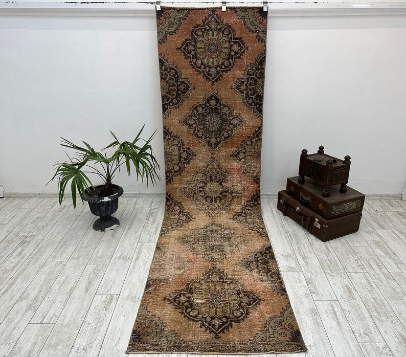Puede incluir: Una alfombra larga y rectangular con un aspecto descolorido y vintage. La alfombra presenta un patr&oacute;n repetitivo de intrincados dise&ntilde;os florales en tonos marrones, rosas y crema. La alfombra es perfecta para a&ntilde;adir un toque de estilo bohemio a cualquier habitaci&oacute;n.