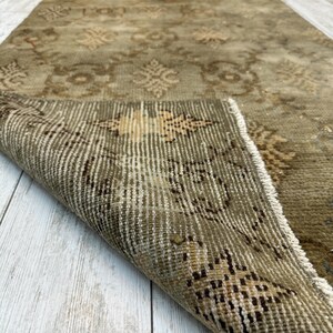 May include: A long, rectangular runner rug in muted olive green and beige tones. The rug features a repeating geometric and floral pattern across its surface. One corner of the rug is folded over, revealing the woven texture and the underside construction.