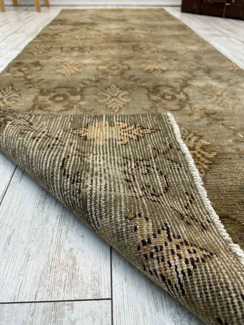 May include: A long, rectangular runner rug in muted olive green and beige tones. The rug features a repeating geometric and floral pattern across its surface. One corner of the rug is folded over, revealing the woven texture and the underside construction.