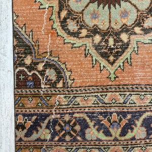 May include: A vintage rug with a faded orange background and intricate floral patterns in shades of brown, green, and blue. The rug has a fringed edge and shows signs of wear and tear.