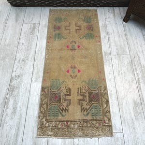Puede incluir: Una alfombra rectangular tipo pasillero con una base de color beige desvanecido y patrones geométricos simétricos en tonos verde, marrón y rosa apagados. El diseño incluye motivos florales estilizados y detalles de bordes complejos y desgastados.