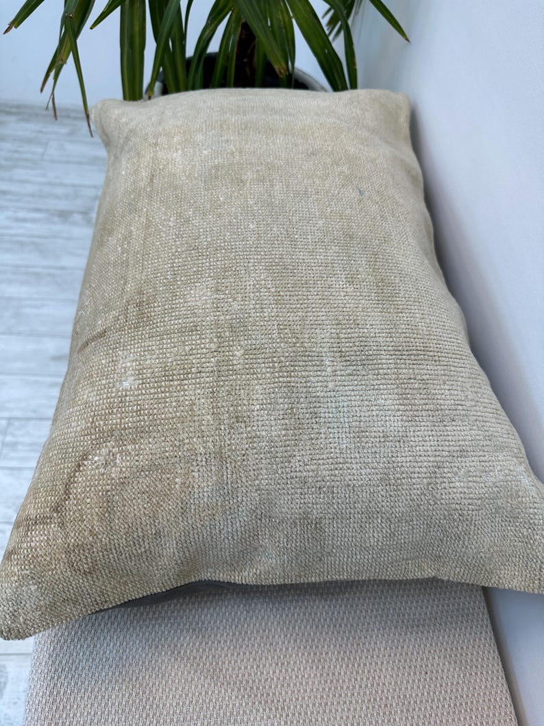 May include: A rectangular throw cushion featuring a textured, woven fabric in a neutral beige and tan colour palette. The surface displays a dense, consistent weave pattern with subtle variations in tone, creating a natural, earthy appearance.