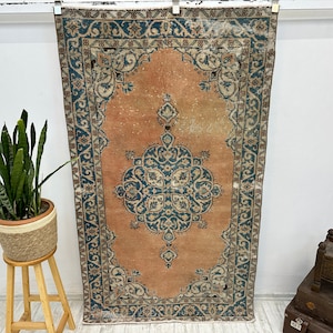 May include: A vintage rug with a peach-colored background and a blue and cream floral pattern. The rug has a fringed edge and is hanging on a white wall.