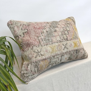 May include: A rectangular throw pillow featuring a woven textile cover with a faded, distressed pattern in muted shades of dusty pink, soft gray, cream, and mustard yellow, displaying geometric shapes and intricate, worn-in decorative motifs.