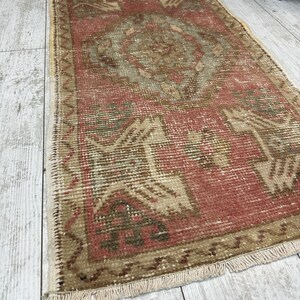 Puede incluir: Una alfombra rectangular vintage con aspecto desgastado. La alfombra presenta un dise&ntilde;o central de medall&oacute;n en tonos rojos, beige y marr&oacute;n. Los bordes est&aacute;n bordeados con una franja, y la alfombra est&aacute; colocada sobre un suelo de madera clara.