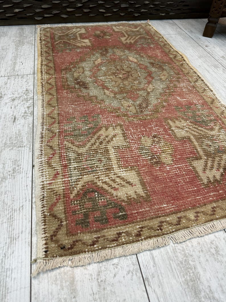 Puede incluir: Una alfombra rectangular vintage con aspecto desgastado. La alfombra presenta un dise&ntilde;o central de medall&oacute;n en tonos rojos, beige y marr&oacute;n. Los bordes est&aacute;n bordeados con una franja, y la alfombra est&aacute; colocada sobre un suelo de madera clara.