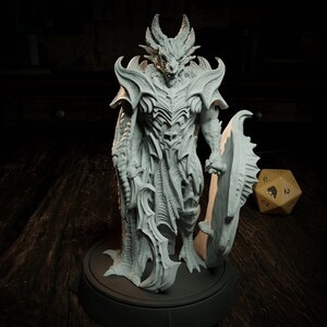 Peut inclure: Une figurine miniature blanche et détaillée d'une créature ressemblant à un dragon portant une armure et tenant une épée et un bouclier. La figurine est debout sur une base ronde.