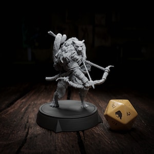 Peut inclure: Une figurine miniature grise et détaillée d'une créature anthropomorphe poilue tenant un arc et une flèche. La créature est debout sur une base circulaire noire. Un dé jaune à 20 faces avec un symbole d'ours est au premier plan.