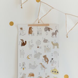 May include: A white poster with a colorful alphabet featuring animals and objects. Each letter is paired with an illustration, such as a fox for F, a goose for G, and a hot air balloon for H. The poster is hanging on a white wall with a string of gold and white flags.