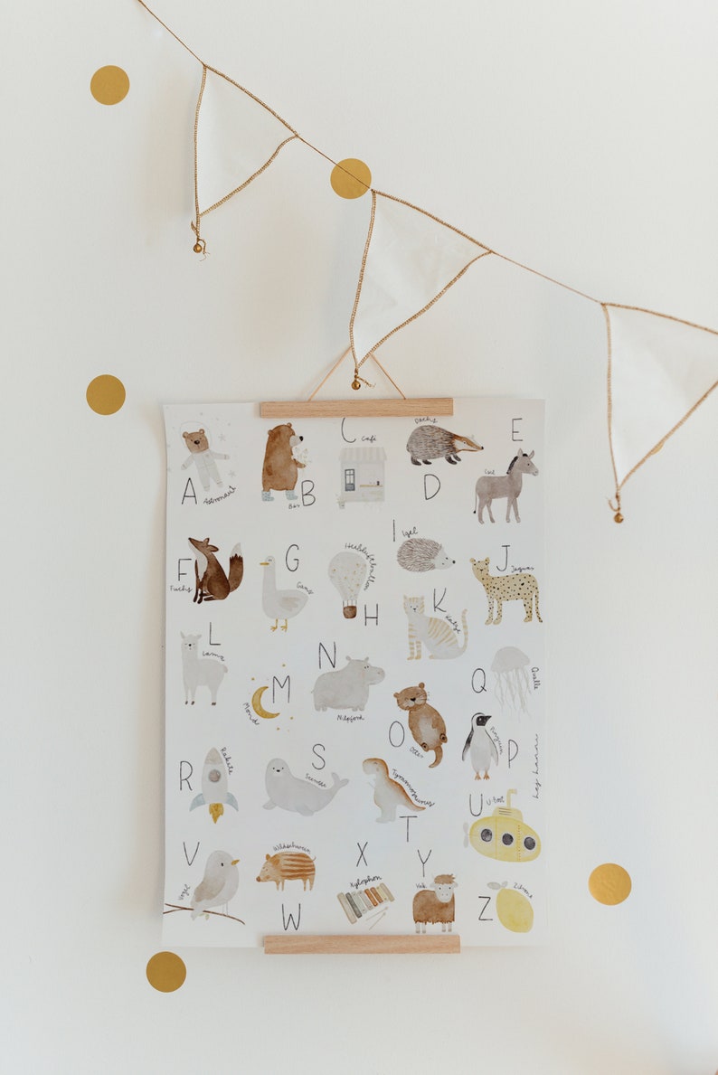 May include: A white poster with a colorful alphabet featuring animals and objects. Each letter is paired with an illustration, such as a fox for F, a goose for G, and a hot air balloon for H. The poster is hanging on a white wall with a string of gold and white flags.