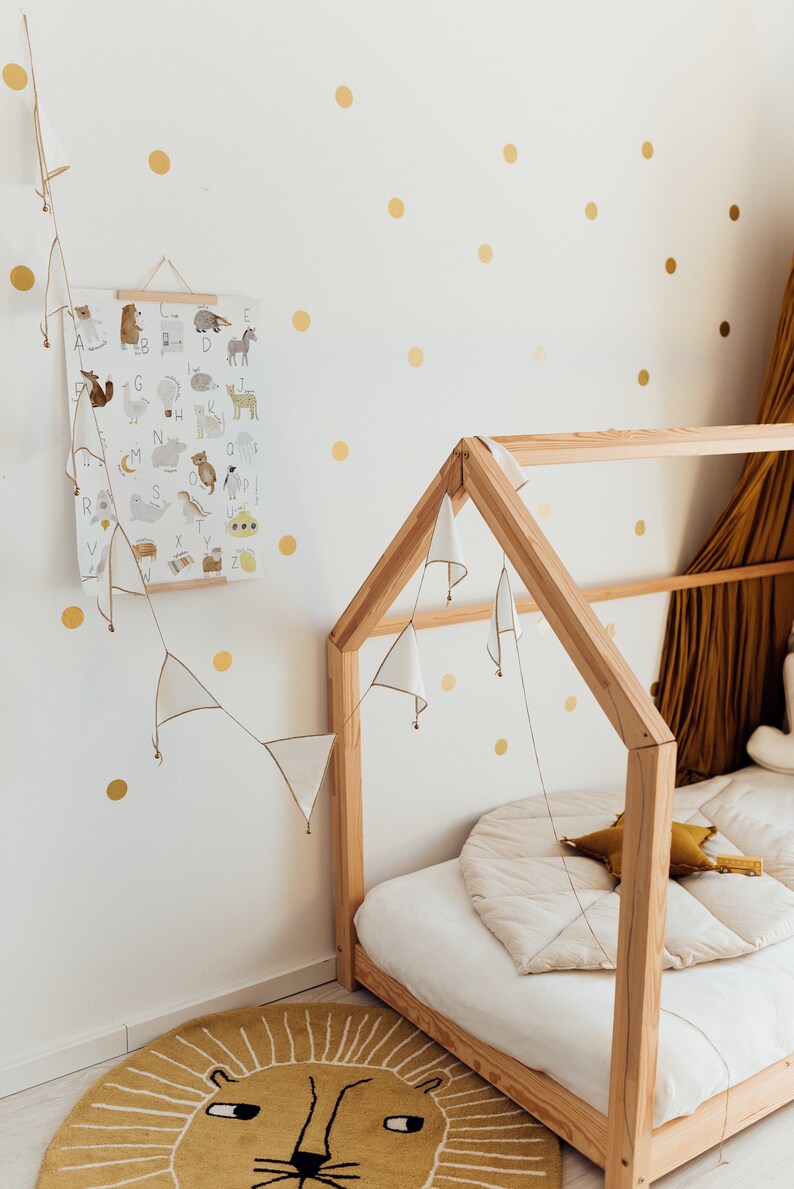 May include: A wooden house-shaped bed frame with a white mattress and a white and gold patterned rug. The wall behind the bed is decorated with gold polka dots and a white poster with an alphabet and animal illustrations.