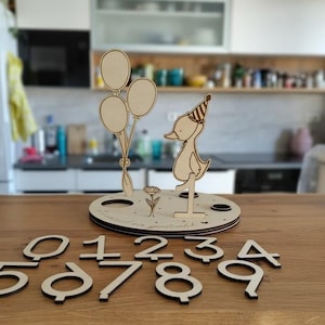 Könnte beinhalten: Geburtstagsdekoration aus Holz mit einer Ente mit Partyhut, Luftballons und einer Blume. Enthält separate Holznummern 0-9. Die Basis hat ein kreisförmiges Design mit Aussparungen für die Ente, Blume und Luftballons.