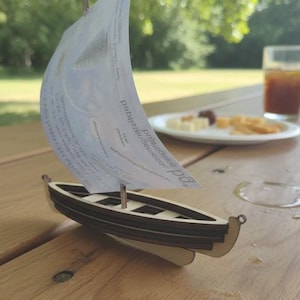 May include: A small wooden sailboat with a paper sail, resting on a wooden picnic table. The boat has a dark brown hull and a light-colored base. A glass of iced tea and a plate of snacks are in the background.