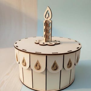 May include: A wooden birthday cake-shaped box with a candle on top. The box is white with brown trim and decorative tear-drop shapes around the base. The candle is also made of wood and has a flame.