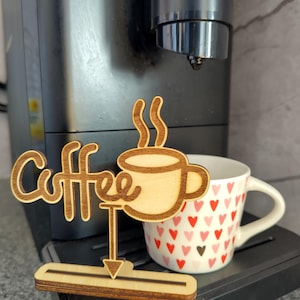 Puede incluir: Letrero de madera con la palabra "Coffee" y un diseño de taza de café. El letrero está colocado frente a una cafetera y una taza con un patrón de corazones.