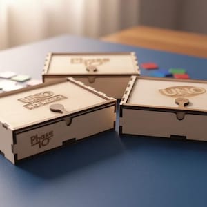 Könnte beinhalten: Drei Holzspielboxen mit gravierten Deckeln. Die Boxen sind hellbraun mit dunkelbraunen Verzierungen und zeigen die Logos "UNO" und "Phase 10". Die Boxen stehen auf einer blauen Oberfläche.