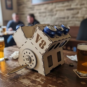 May include: A wooden V8 engine-shaped beer can holder with a gear and handle. The holder is filled with blue and silver cans. Two glasses of beer are on the table. The word "V8" is visible on the holder.