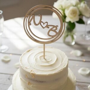 Op de afbeelding: Een bruidstaart met twee lagen, witte glazuur en gouden bladaccenten. Een houten taarttopper met de letters "W" en "B" in een cirkel staat bovenop de taart. Een boeket witte bloemen staat op de achtergrond.