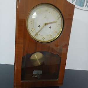 May include: A vintage wooden wall clock with a light-coloured face and gold-tone hands and numerals. The clock has a pendulum visible through a glass panel. The clock face has the word "Junghans" printed on it.